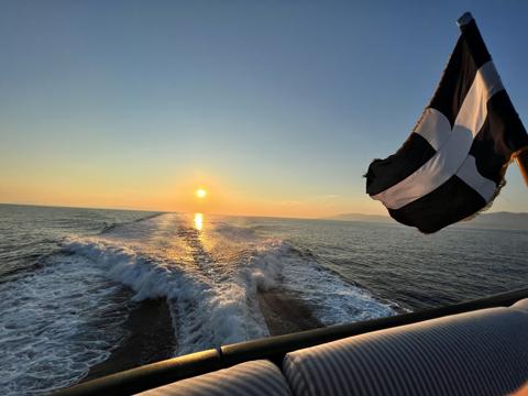 Sunset from the stern of a boat in the Cyclades: the wake running back toward the low sun, a cross flag snapping in the wind, hills on the far horizon