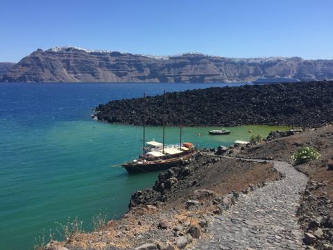 A wooden caique moored in green water beside black lava rock, the caldera cliff and its white towns behind