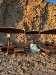 Thatched umbrellas and loungers on the sand below an ochre cliff at golden hour, Milos