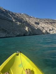 The bow of a yellow kayak on clear turquoise water below pale cliffs, Milos