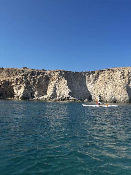 White volcanic cliffs above deep teal water, a kayak crossing below them, Milos
