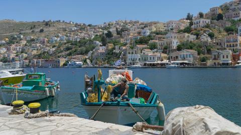 A fisherman working from his boat in Symi harbour, with the island's pastel houses behind him