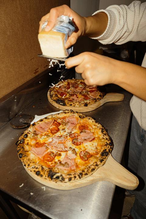 Two blistered pizzas on wooden peels at the pass, hard cheese grated over the top
