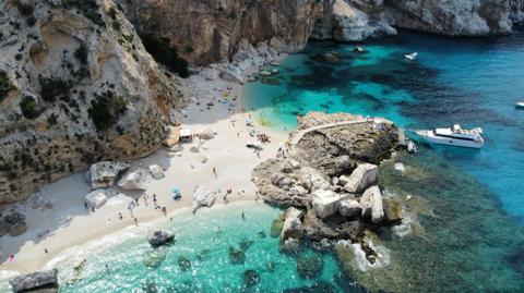 A cliff-backed cove with a pale pebble beach, boats anchored off it