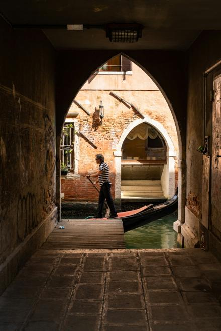 A gondolier poling past, framed in a dark covered passage that opens onto a canal