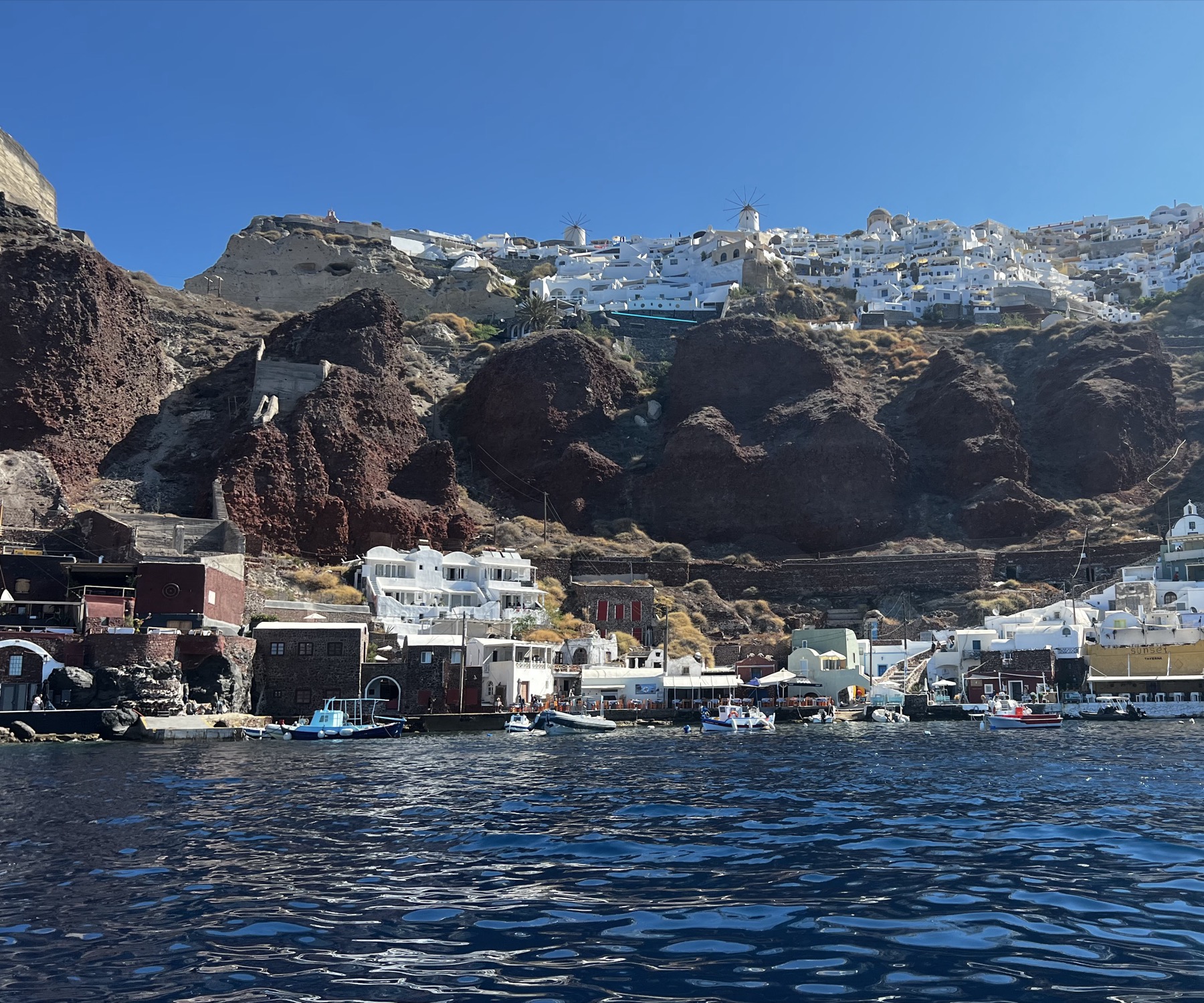 Oia on the cliff above Ammoudi Bay, seen from the water: windmills on the ridge, red volcanic rock, tavernas and moored boats at the waterline
